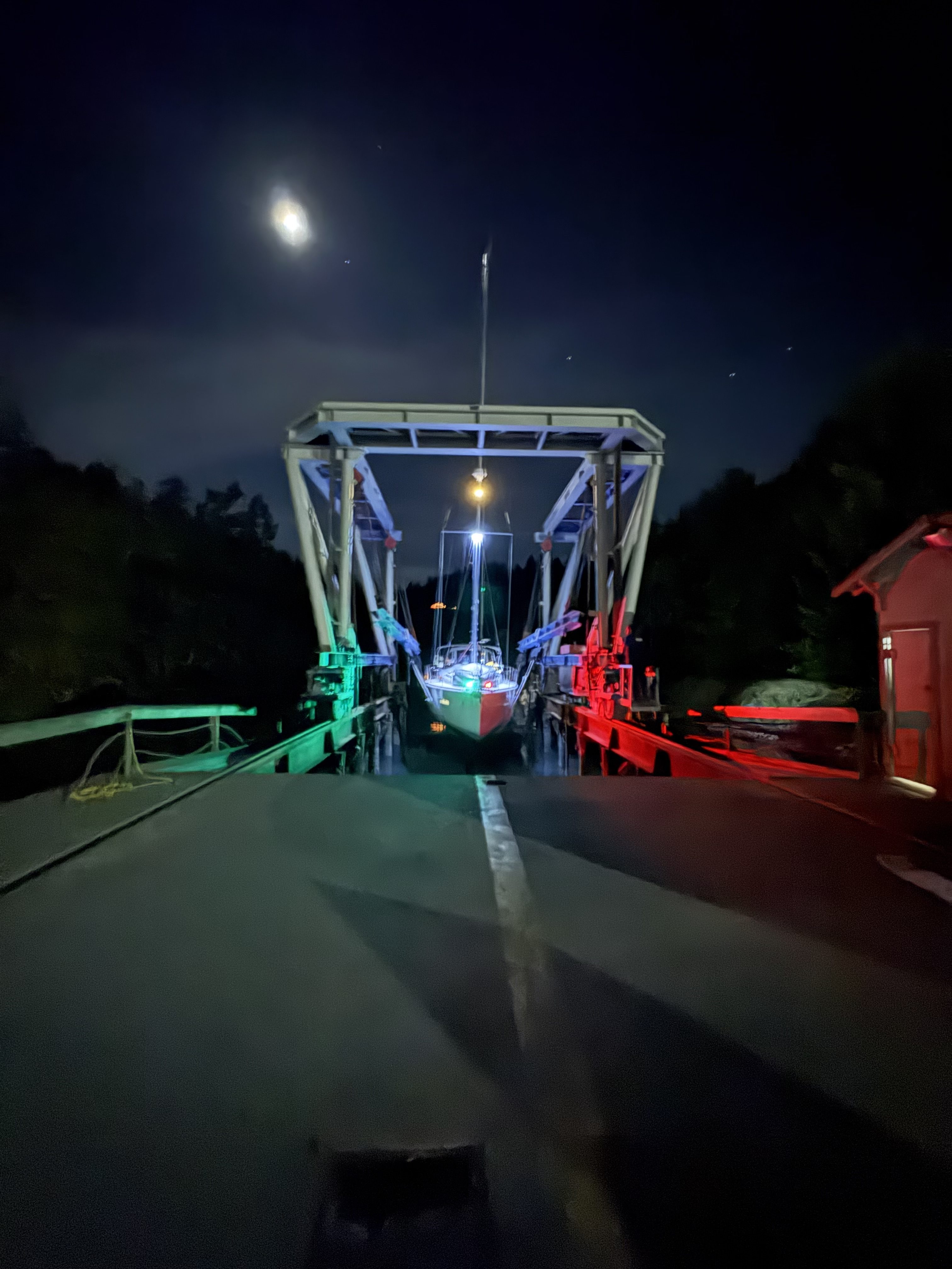 The travel lift at night with navigation lights lit and a sailboat in the slings at Essential Shipyard